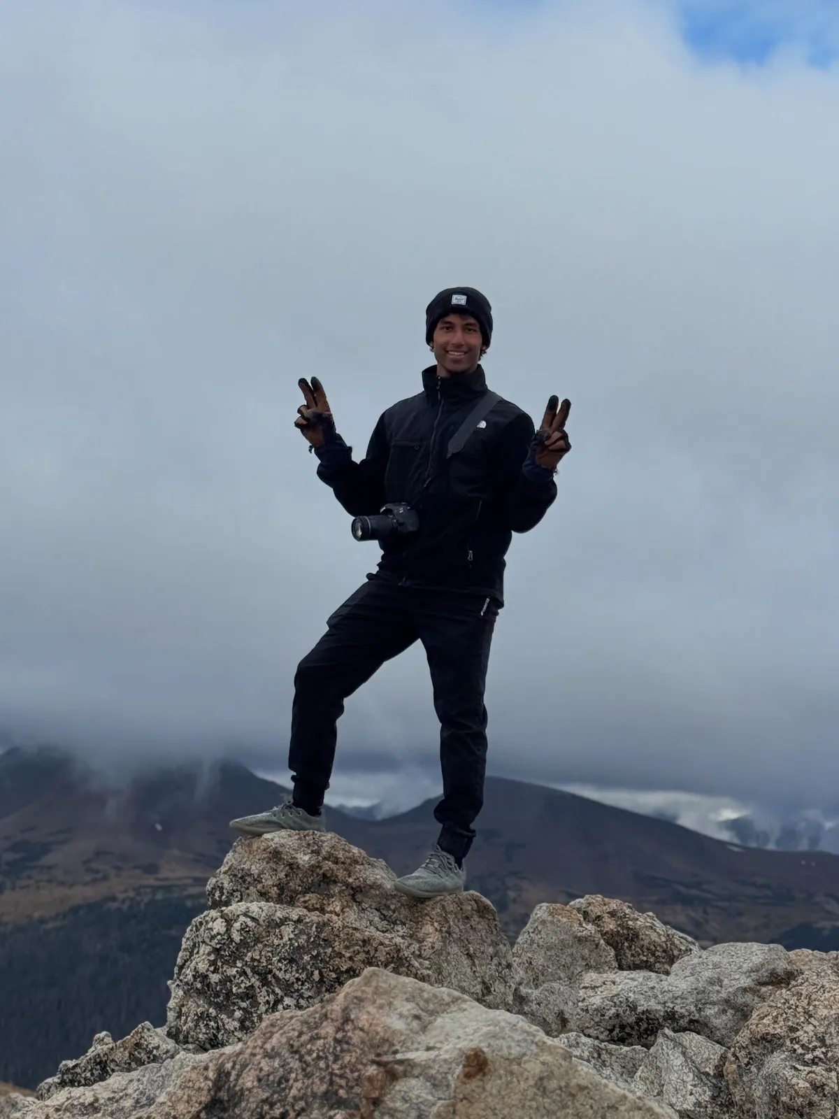 Sai standing on a mountain peak, dressed in black, holding a camera at his chest